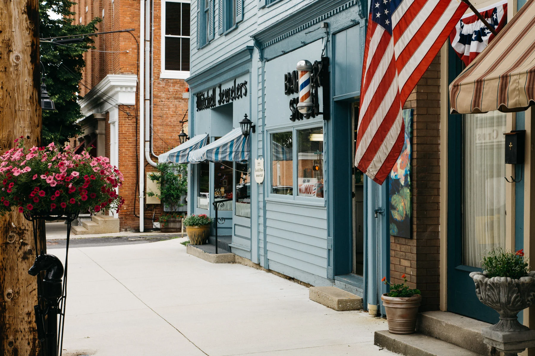 An American flag and flower baskets outside shops on a Bedford sidewalk