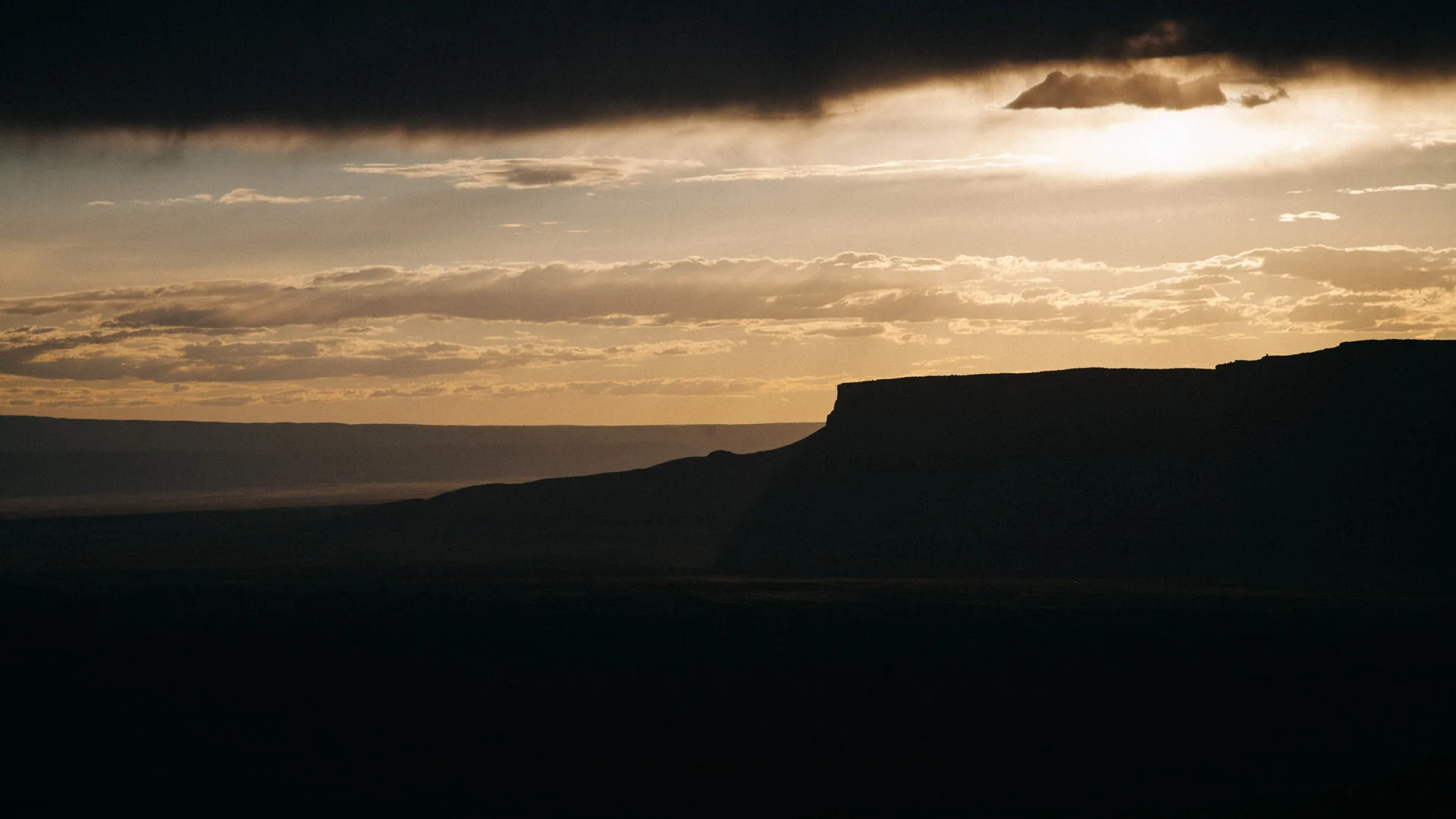 The sun breaking through dark clouds over a silhouetted mesa