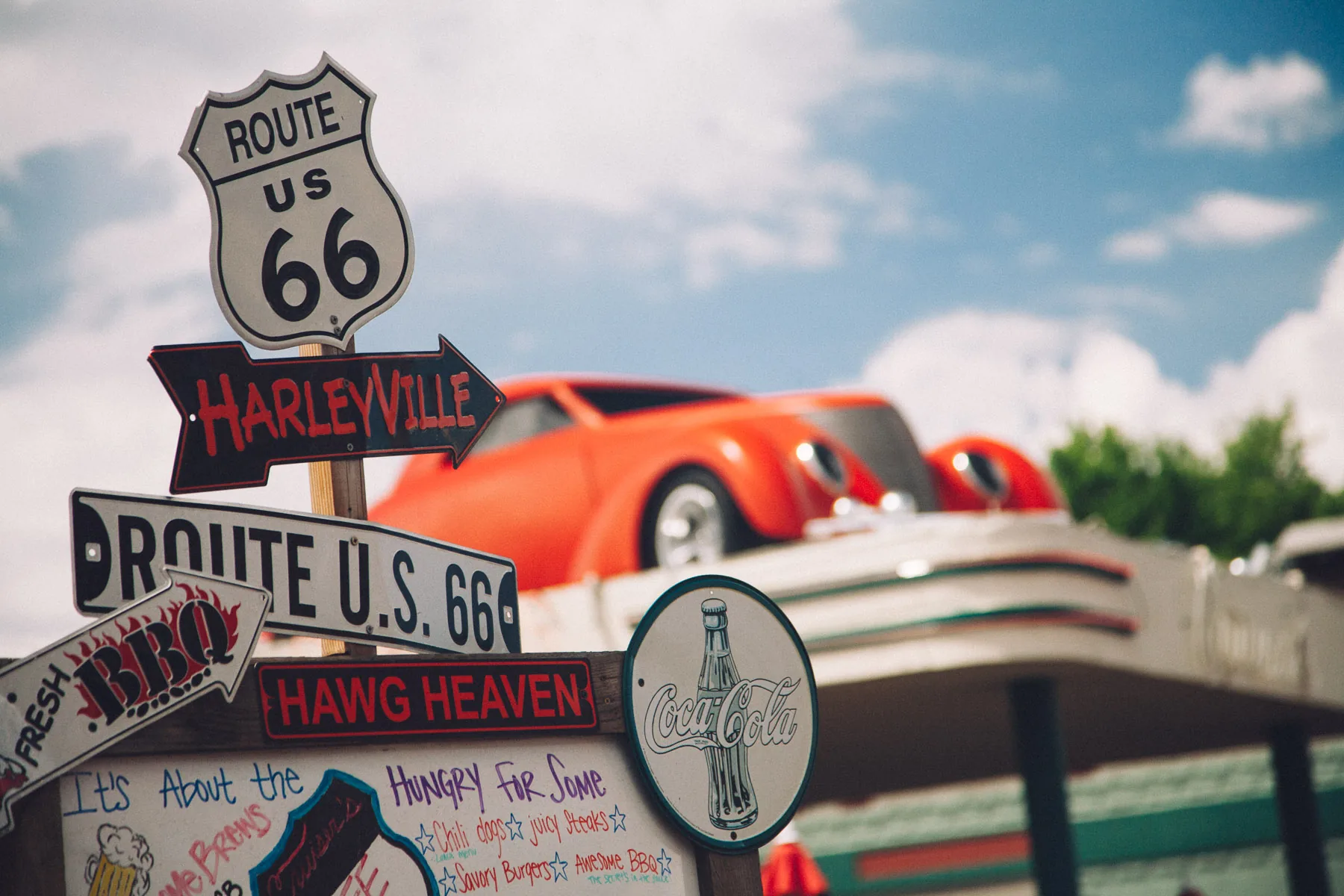 Route 66 and Harleyville signs below a red hot rod on a roof
