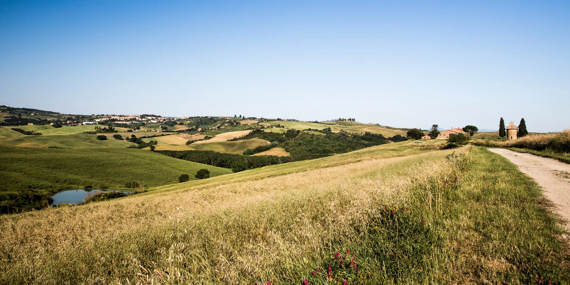Dirt road along a grassy ridge overlooking a pond and distant farms