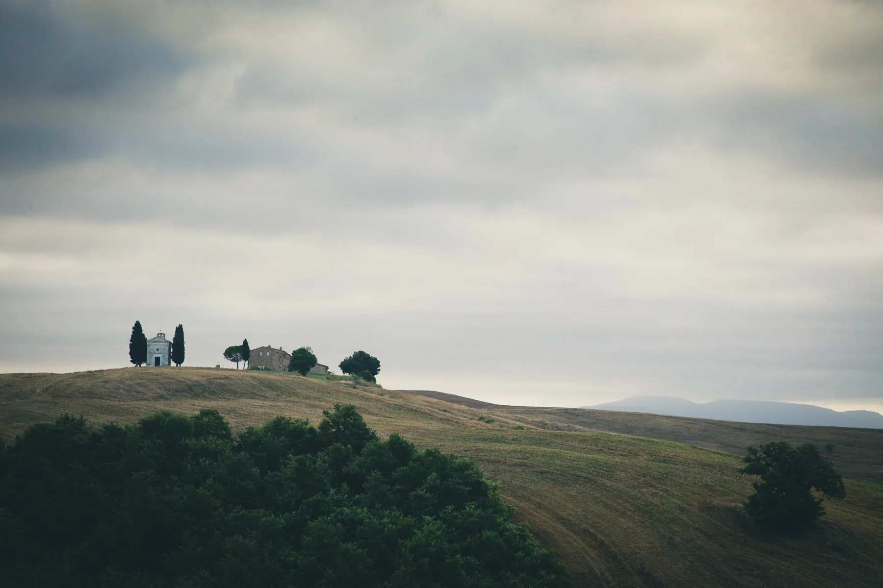 Chapel and farmhouse on a distant ridge above dark woods under grey cloud